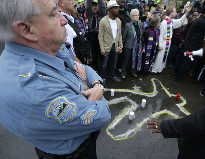 Un agente de polic&iacute;a de Ferguson, Missouri, permanece de pie en la calle ante la comisar&iacute;a de polic&iacute;a cerca de una silueta dibujada con tiza en recuerdo de Michael Brown, el 13 de octubre de 2014. (Foto AP/Charles Rex Arbogast)