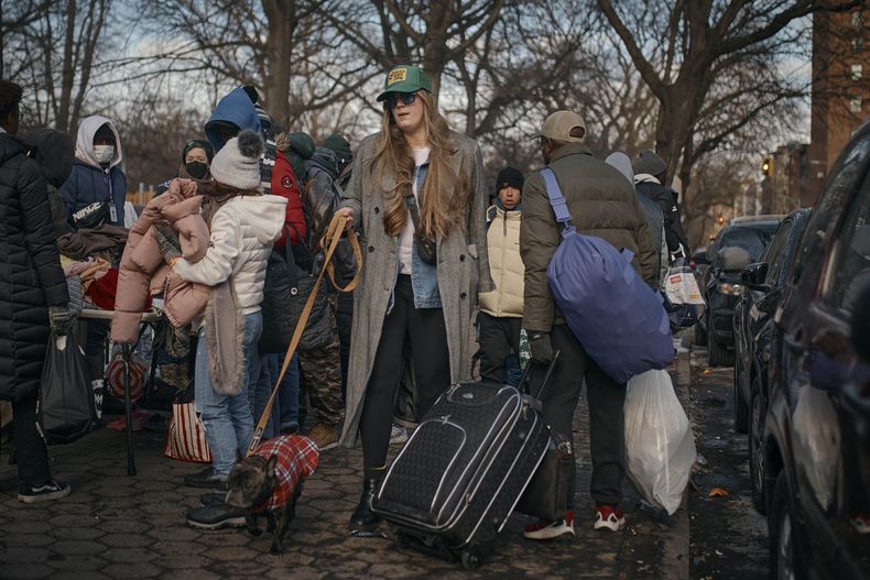 Una mujer pasea a su perro mientras migrantes recogen ropa distribuida por grupos de ayuda cerca del Centro de Asistencia al Migrante en la Escuela Primaria Santa Brígida, el sábado 20 de enero de 2024, en Nueva York. (AP Foto/Andres Kudacki)