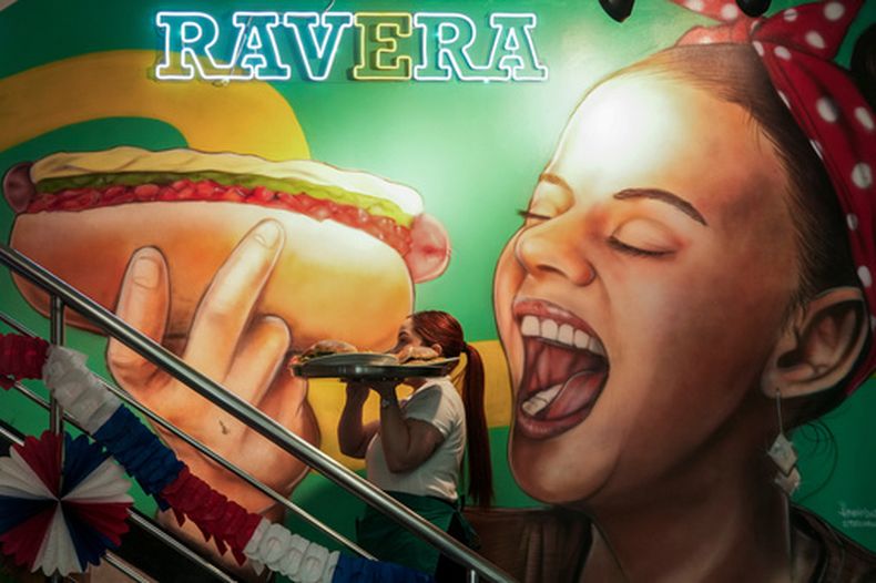 Una camarera lleva una bandeja con completos -los tradicionales perros calientes chilenos con puré de aguacate, tomate picado y mayonesa- pasando frente a un mural dedicado a este plato en el restaurante Ravera, en el centro de Santiago de Chile, el jueves 10 de septiembre de 2026. (AP Foto/Esteban Félix)