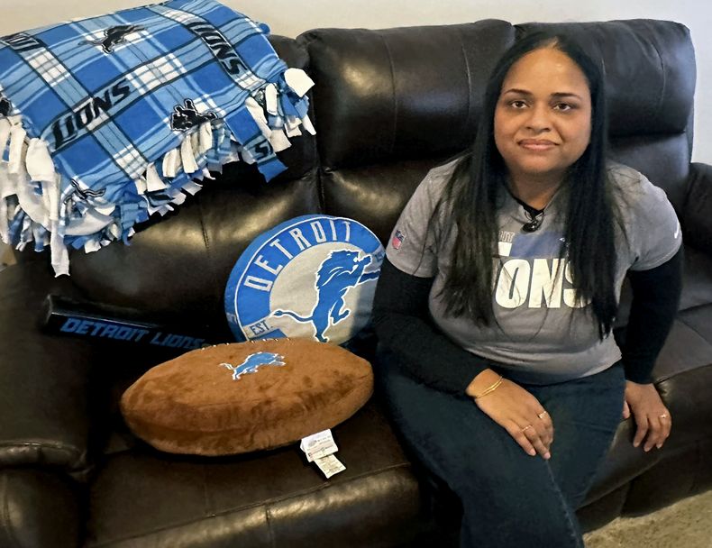 Sweta Patel sentada en su hogaren Canton, Michagan, con almohadas de los Lions de Detroit, un mantel y un megáfono el jueves 18 de enero de 2024. Sweta Patel sits in her Canton, Mich., home with Detroit Lions pillows, a blanket and speaker Thursday, Jan. 18, 2024. (AP Foto/Larry Lage)