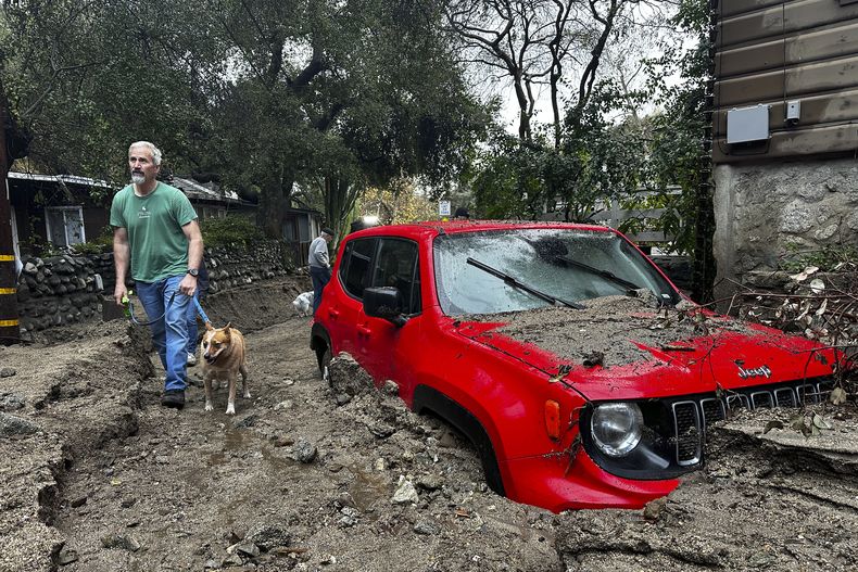 Un residente y su perro caminan junto a un vehículo parcialmente sumergido en lodo tras una tormenta, el viernes 14 de febrero de 2025, en Sierra Madre, California. (AP Foto/Eugene Garcia)