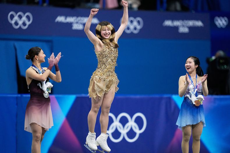 La estadounidense Alysa Liu, que ganó el oro en el patinaje artístico, salta en el centro de la foto flanqueada por las japonesas Kaori Sakamoto (plata) y Ami Nakai (bronce) en el podio el jueves 19 de febrero del 2026. (AP Foto/Natacha Pisarenko)