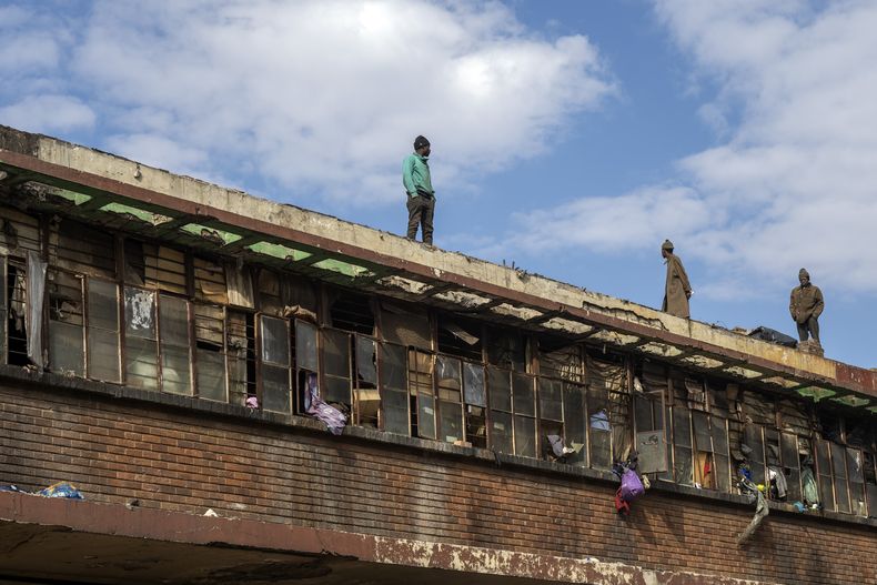 Ocupantes ilegales de viviendas de pie en un techo viendo el sitio donde se produjo uno de los incendios más mortíferos de Sudáfrica, en la capital Johannesburgo, el viernes 1 de septiembre de 2023. (AP Foto/Jerome Delay)