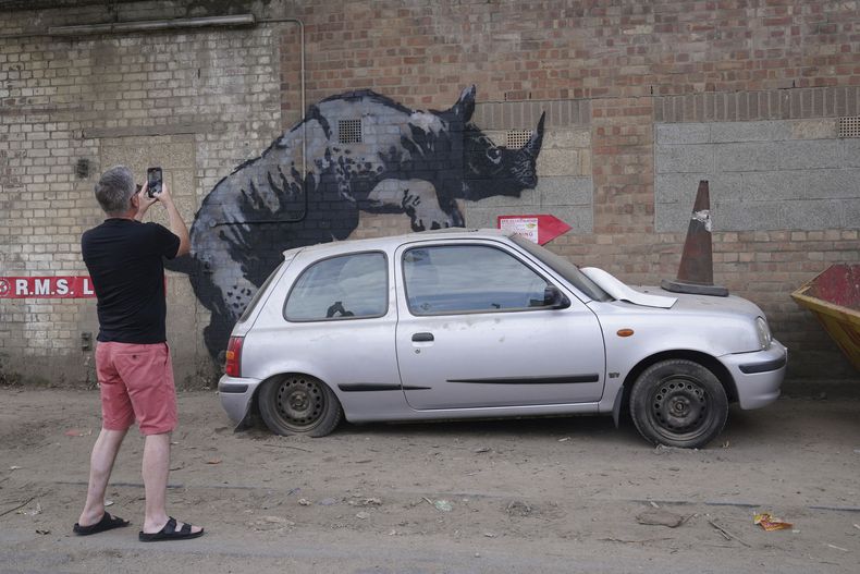 Un hombre fotografía una nueva obra de arte de Banksy, que representa un rinoceronte que parece subirse a un automóvil, la octava obra de arte de su colección de temática animal, en Westmoor Street en Charlton, sureste de Londres, el lunes 12 de agosto de 2024. (Lucy North/PA via AP)
