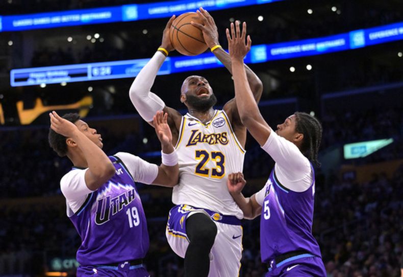 LeBron James (23), alero de los Lakers de Los Ángeles, es defendido por Ace Bailey, a la izquierda, y el alero Cody Williams, de los Jazz de Utah, mientras conduce hacia la canasta durante la primera mitad de un juego de baloncesto de la NBA, el domingo 12 de abril de 2026, en Los Ángeles. (Foto AP/Jayne Kamin-Oncea)