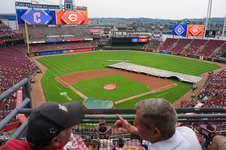 Fanáticos platican mientas que una cuadrilla de trabajadores de los Rojos de Cincinnati cubren el diamante durante el retraso por lluvia previo al partido contra los Guardianes de Cleveland, el lunes 27 de julio de 2026, en Cincinnati. (AP Foto/Kareem Elgazzar)