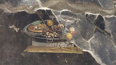 Esta imagen proporcionada el martes 27 de junio de 2023 por el Sitio Arqueológico de Pompeya muestra la pared de una antigua casa pompeyana con un fresco que representa una mesa con comida. El fresco fue encontrado en el atrio de una casa en Insula 10 de Regio IX en una excavación, a la que se anexó una panadería, ya explorada parcialmente entre 1888 y 1891 y cuyas investigaciones se reanudaron en enero pasado. (Sitio Arqueológico de Pompeya vía AP, ho)