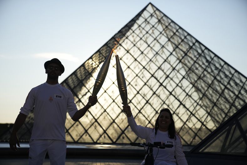 ARCHIVO - El fotógrafo frances JR, izquierda, y la atleta Sandra Laoura portan la antorcha olímpica en la explanada del Museo Louvre el domingo 14 de julio, en París. La propia ciudad será una de las protagonistas de los Juegos Olímpicos de 2024. Es por eso que la Torre Eiffel y el río Sena aparecen tan prominentemente como Simone Biles o Katie Ledecky en los anuncios de NBC. (Foto AP/Christophe Ena, Archivo)