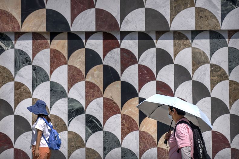 Una persona utiliza un paraguas para protegerse del calor, mientras que otra lo hace con un sombrero, mientras caminan por el exterior de un centro comercial en un día de mucho calor en Beijing, el 24 de junio de 2023. (AP Foto/Mark Schiefelbein)
