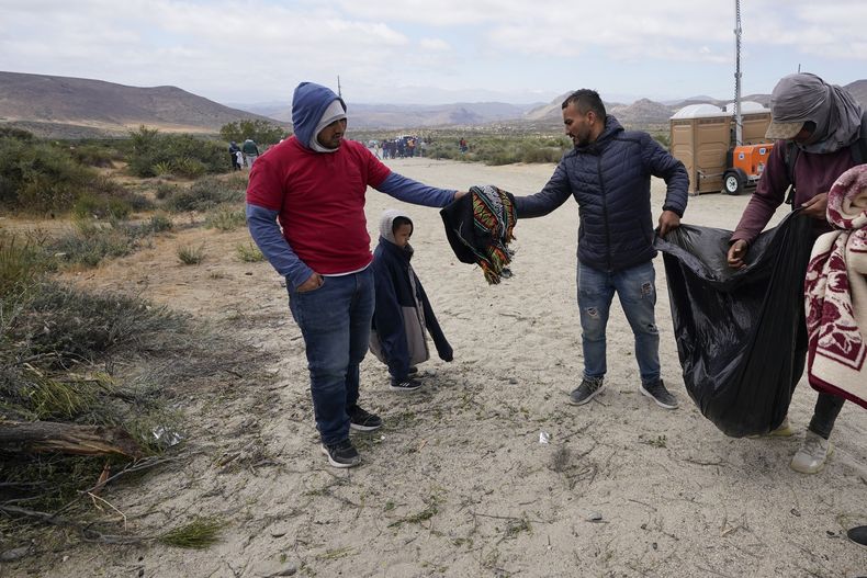 Un migrante colombiano, al centro, le entrega una frazada a un padre y su hijo, también colombianos, mientras el grupo espera a solicitar asilo después de cruzar la frontera, el miércoles 10 de mayo de 2023, cerca de Jacumba, California. El grupo había estado acampando del otro lado de la frontera durante días, a la espera de pedir asilo en Estados Unidos. Mientras los miembros del grupo avanzan al frente para ser llevados a vagonetas, entregan toda su ropa gruesa a los que podrían permanecer otra noche en el campamento. (AP Foto/Gregory Bull)