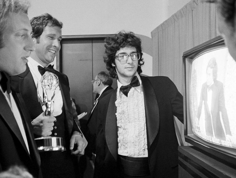 ARCHIVO - Chevy Chase, miembro del elenco de Saturday Night Live, sostiene su premio Emmy mientras observa un monitor que muestra al productor Lorne Michaels aceptando otro Emmy por el programa, mientras los escritores Alan Zweibel, izquierda, y Tom Davis, derecha, observan durante la 28ª edición de los premios Primetime Emmy en Los Ángeles el 17 de mayo de 1976. (Foto AP/Reed Saxon, archivo)