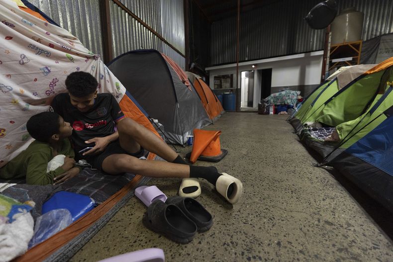 Mickel Rodríguez, de 12 años, de Venezuela, a la izquierda, mira videos en el celular de un amigo de la familia que viaja con su familia mientras descansan en su tienda de un refugio de migrantes en Tijuana, México, el domingo 2 de febrero de 2025. (AP Foto/Gregory Bull)