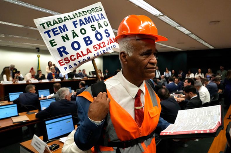 El legislador y pastor Sargento Isidorio sostiene un cartel en apoyo al fin de la semana laboral de seis días en Brasil, que dice en portugués: Los trabajadores tienen familias y no son robots. ¡Ya debemos implementar la jornada laboral 5x2!, durante una sesión especial del comité que analizaba la propuesta en la Cámara de Diputados, el miércoles 27 de mayo de 2026, en Brasilia, Brasil. (AP Foto/Eraldo Peres)