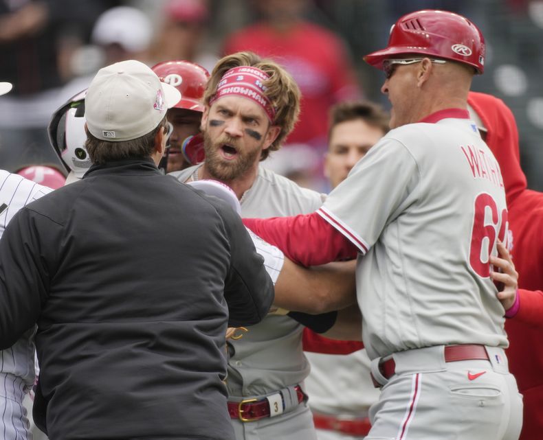 Bryce Harper, centro, de los Filis de Filadelfia, es contenido por el umpire de primera, Ben May, izquierda, y el coach de tercera de los Filis, Dusty Wathan, después de reaccionar a una burla del relevista de los Rockies de Colorado, Jake Bird, en la séptima entrada del juego de béisbol, el domingo 14 de mayo de 2023, en Denver. (AP Foto/David Zalubowski)