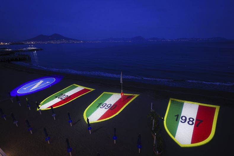 El escudo del Napoli y tres Scudettos referentes a sus dos títulos anteriores y al que podría conseguir, se proyectan en la playa de Posillipo, el jueves 27 de abril de 2023 (Alessandro Garofalo/LaPresse via AP)