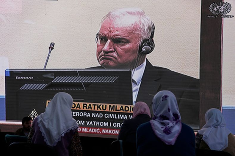 ARCHIVO – Mujeres de Srebrenica miran una transmisión en vivo del Tribunal Penal Internacional para la antigua Yugoslavia en La Haya para conocer el veredicto del jefe militar serbobosnio Ratko Mladic, en pantalla, en el cementerio memorial en Potocari cerca de Srebrenica, en el este de Bosnia, el martes 8 de junio de 2021.(AP Foto/Darko Bandic, Archivo)