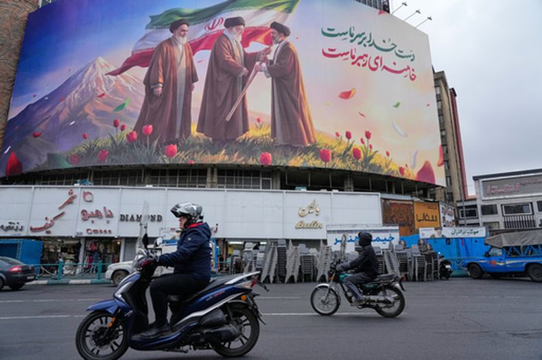 ARCHIVO - Motos pasan por delante de un cartel con la imagen del difunto líder supremo de Irán, el ayatolá Ali Jamenei (centro), entregando la bandera del país a su hijo y sucesor, el ayatolá Moytabá Jamenei (derecha), con el fallecido líder revolucionario, el ayatolá Ruhollah Jomeini a la izquieda, en una plaza en el centro de Teherán, Irán, el 10 de marzo de 2026. (AP Foto/Vahid Salemi, archivo)