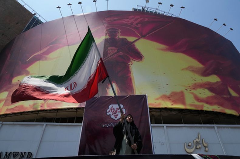 Una mujer ondea una bandera iraní junto a una valla publicitaria que muestra la imagen de un hombre con una camiseta que trae el rostro del difunto líder supremo iraní, el ayatolá Alí Jamenei --quien murió durante un ataque conjunto de Estados Unidos e Israel el 28 de febrero--, con palabras que invocan a los vengadores del imán Hussein, uno de los santos más venerados del islam chií, el sábado 1 de agosto de 2026, en una plaza del centro de Teherán, Irán. (AP Foto/Vahid Salemi)