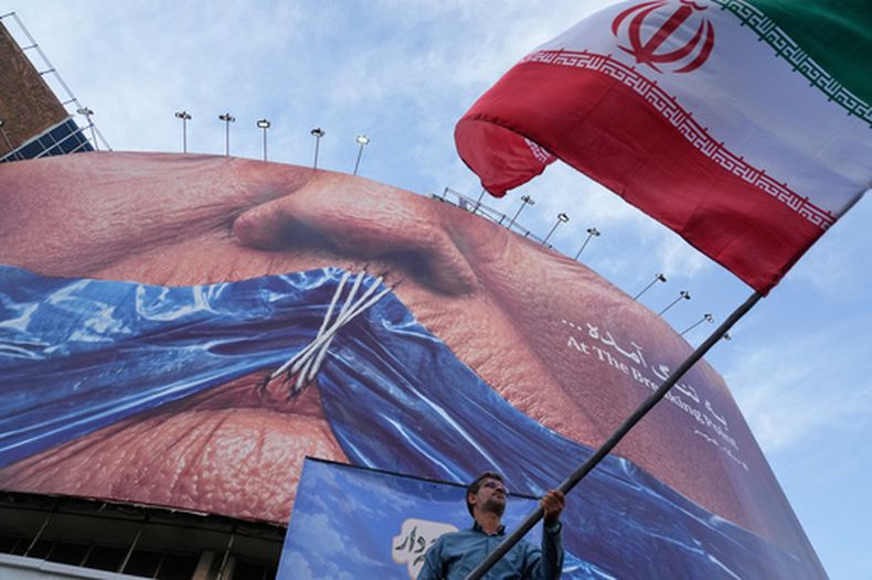 Un hombre ondea una bandera iraní para una campaña progubernamental bajo un cartel que muestra el estrecho de Ormuz y los labios del presidente de Estados Unidos, Donald Trump, cosidos, en una plaza en el centro de Teherán, Irán, el 6 de mayo de 2026. (AP Foto/Vahid Salemi)