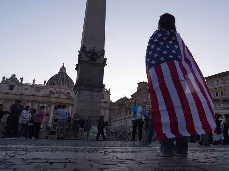Andrea Gallardo, de 20 años, quien vive en Texas, observa envuelta en una bandera estadounidense después de que el papa León XIV salió al balcón de la Basílica de San Pedro tras ser elegido, el jueves 8 de mayo de 2025, en el Vaticano. (AP Foto/Paolo Santalucia)