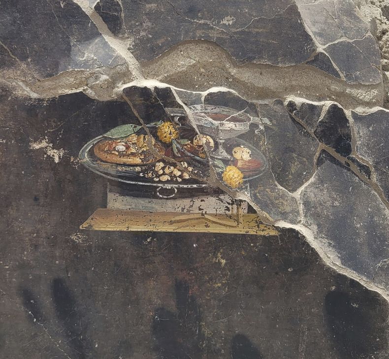 Esta imagen proporcionada el martes 27 de junio de 2023 por el Sitio Arqueológico de Pompeya muestra la pared de una antigua casa pompeyana con un fresco que representa una mesa con comida. El fresco fue encontrado en el atrio de una casa en Insula 10 de Regio IX en una excavación, a la que se anexó una panadería, ya explorada parcialmente entre 1888 y 1891 y cuyas investigaciones se reanudaron en enero pasado. (Sitio Arqueológico de Pompeya vía AP, ho)