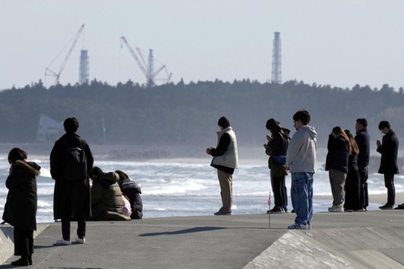 Gente guarda un momento de silencio a las 2:46 de la tarde, el momento en el que se produjo un sismo, con la central nuclear de Fukushima Daiichi al fondo, en Namie, en la prefectura de Fukushima, en el norte de Japón, mientras el país conmemora el 15 aniversario del enorme terremoto, tsunami y desastre nuclear. (Kyodo News via AP)