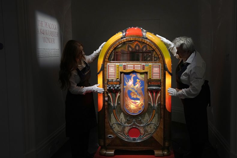 Empleados de Sothebys colocan una rocola de diseño Peacock Wurlitzer Modelo 850 de Paul Fuller, estadounidense, de cerca de 1941, en las salas de subastas de Sothebys en Londres, el jueves 3 de agosto de 2023. Más de 1.400 artículos personales de Freddie Mercury, incluidos sus extravagantes vestuarios, borradores escritos a mano de Bohemian Rhapsody y el piano de media cola que usó para componer grandes éxitos de Queen, se exhibirán en la casa de subastas Sothebys Londres antes de su venta. (Foto AP/Kirsty Wigglesworth)