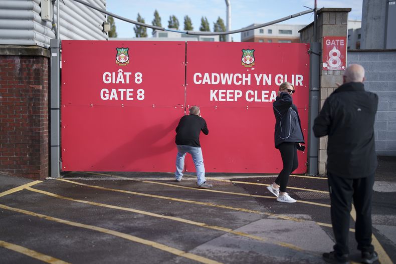 Personas tratan de ver el campo a través de una puerta cerrada en el hipódromo del Wrexham Football Club en Wrexham, Gales, el lunes 7 de octubre de 2024. (Foto AP/Jon Super)