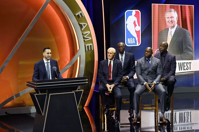 Jonnie West, a la izquierda, habla durante la consagración de su difunto padre Jerry West en el Salón de la Fama del Baloncesto mientras, de izquierda a derecha, Pat Riley, Bob McAdoo, Magic Johnson y James Worthy lo acompañan en el escenario, el domingo 13 de octubre de 2024 en Springfield, Massachusetts. (Foto AP/Jessica Hill)
