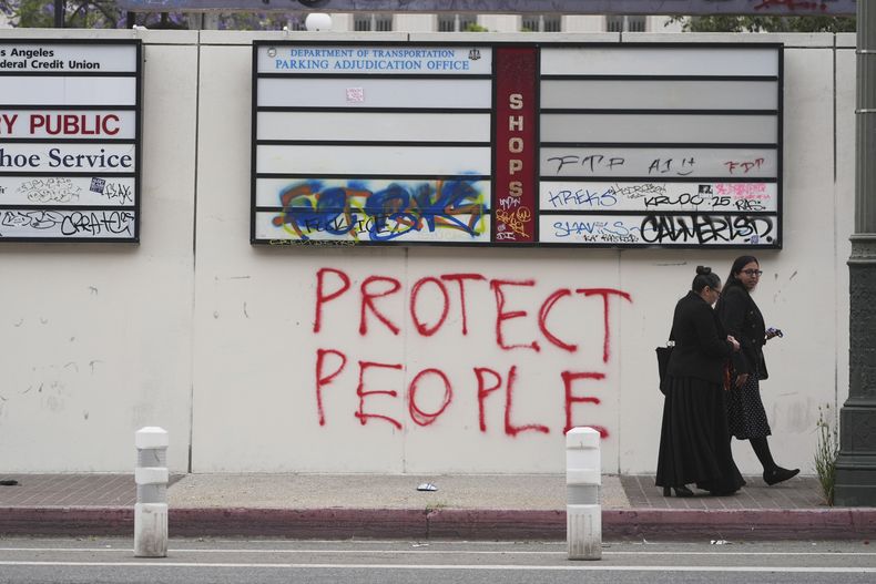 Dos personas pasan por delante de un grafiti con la frase Protejan a la gente, realizado durante las últimas protestas contra las redadas migratorias federales, el 11 de junio de 2025, en Los Ángeles. (AP Foto/Damian Dovarganes)