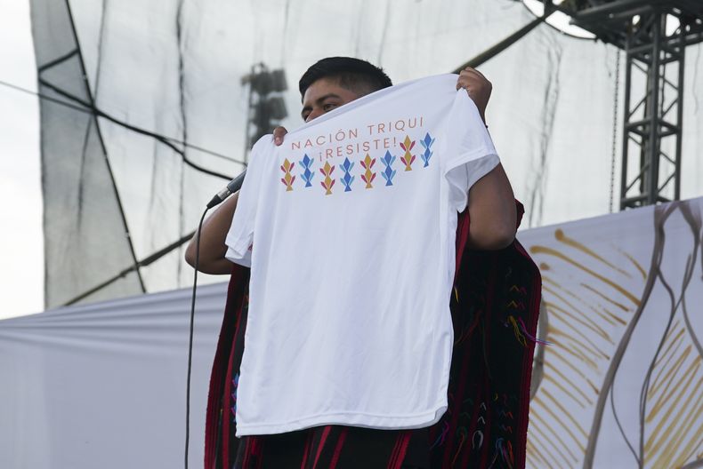 El rapero mexicano-triqui Carlos CGH, vestido con un gabán negro y rojo, una prenda nativa del pueblo triqui en el oeste de Oaxaca, sostiene una camiseta que dice Nación triqui ¡resiste! durante una celebración de los pueblos indígenas en el Zócalo de la Ciudad de México, el martes 6 de agosto de 2024. (Foto AP/Mariana Martínez Barba)