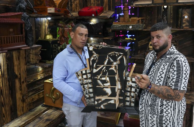 Los artesanos Adrián Morán, izquierda, y Ariel Sánchez, creadores de Humidores Clave, posan con uno de sus humidificadores de cigarros durante la XXV Feria del Habano en el Palacio de Convenciones de La Habana, el lunes 24 de febrero de 2025. (AP Foto/Ramón Espinosa)