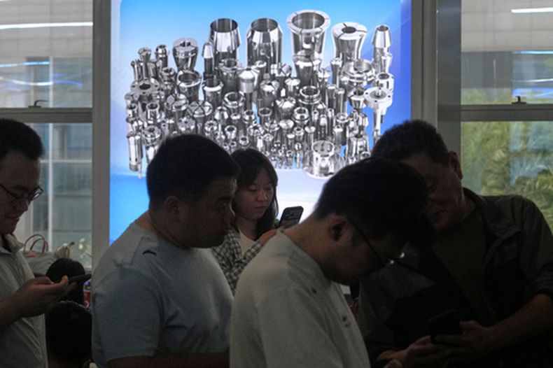 Pasajeros hacen fila para salir de una estación de tren mientras en una pantalla se anuncia una fábrica que elabora piezas para máquinas, en Shanghai, China, el 19 de octubre de 2026. (AP Foto/Andy Wong)