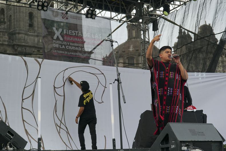 El rapero mexicano-triqui Carlos CGH, vestido con un gabán negro y rojo, una prenda nativa del pueblo triqui en el oeste de Oaxaca, actúa durante una celebración de los pueblos indígenas mientras el artista Alberto Sebastián Bautista Figueroa hace un grafiti en el Zócalo de la Ciudad de México, el martes 6 de agosto de 2024. (Foto AP/Mariana Martínez Barba)