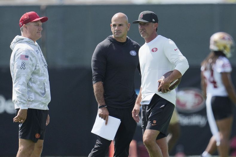 ARCHIVO - El gerente de los 49ers de San Francisco, John Lynch (izquierda), charla con el coordinador defensivo Robert Saleh (medio) y el entrenador en jefe Kyle Shanahan durante un entrenamiento, el 23 de julio de 2025, en Santa Clara, California. (AP Foto/Jeff Chiu)