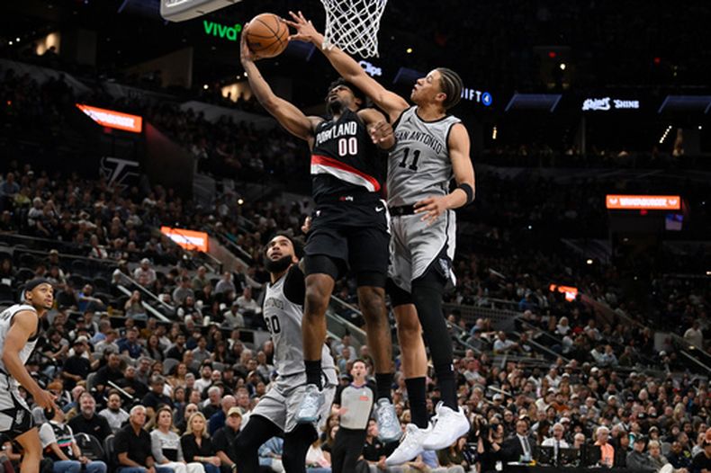 Scoot Henderson (00), guardia de el Trail Blazers de Portland, recibe una falta de Carter Bryant, delantero de los Spurs de San Antonio, durante la primera mitad de un juego de baloncesto de la NBA, el miércoles 8 de abril de 2026, en San Antonio. (Foto AP/Darren Abate)