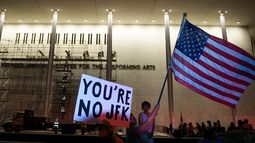 Manifestantes ondean una bandera de Estados Unidos y carteles mientras operarios se preparan para retirar el nombre del presidente Donald Trump del John F. Kennedy Center for the Performing Arts en Washington, el 13 de junio de 2026. (AP Foto/Cliff Owen)