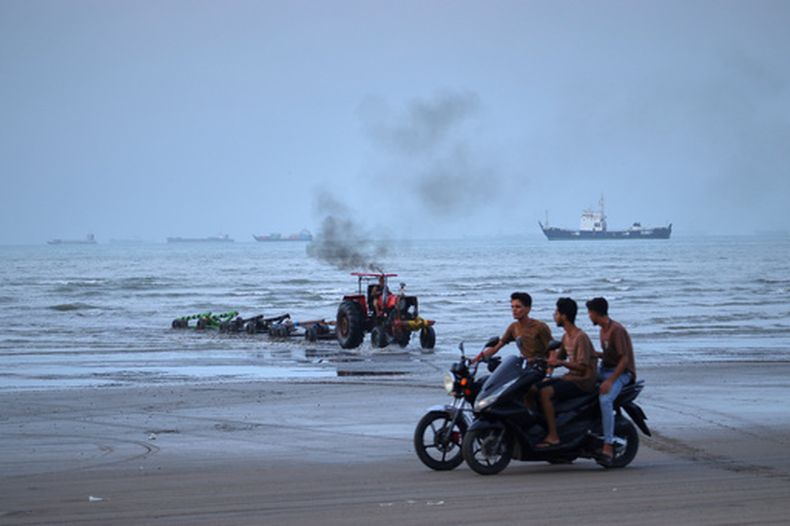 Jóvenes a bordo de motocicletas a lo largo de la orilla mientras un tractor saca equipo del agua y al fondo se pueden ver buques mercantes aparentemente anclados en el estrecho de Ormuz, el lunes 27 de julio de 2026, en Bandar Abbas, Irán. (Razieh Poudat/ISNA vía AP)