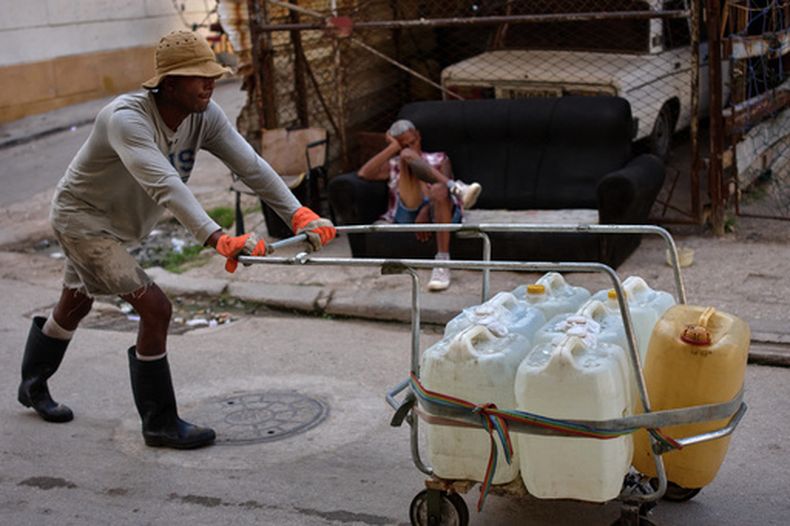 Un hombre empuja un carrito lleno de bidones de agua mientras otro descansa en un sofá en una calle de La Habana, Cuba, el jueves 27 de agosto de 2026. (Foto AP/Ramón Espinosa)