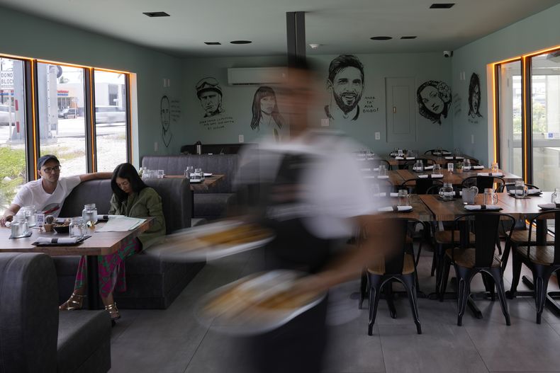 Un camarero lleva una orden frente a un muro en el que aparecen varias celebridades, incluido el astro argentino Lionel Messi, en el Kao Bar & Grill de Hallandale Beach, Florida, el lunes 10 de julio de 2023 (AP Foto/Rebecca Blackwell)