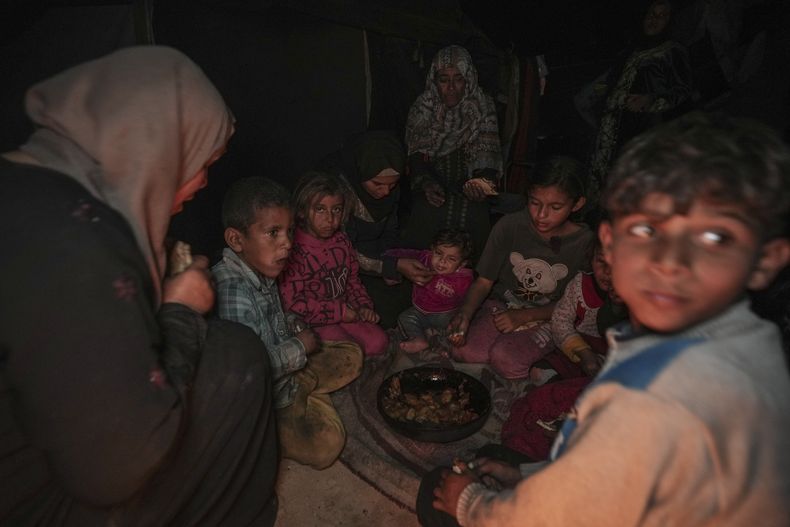 Amani Abu Zarada, cuarta por la izquierda, alimenta a uno de sus hijos con calabacines fritos hechos sobre un fuego hecho con restos de papel y cartón fuera de su tienda de campaña en un campamento de Jan Yunis, Franja de Gaza, jueves 19 de diciembre de 2024. (AP foto/Abdel Kareem Hana)