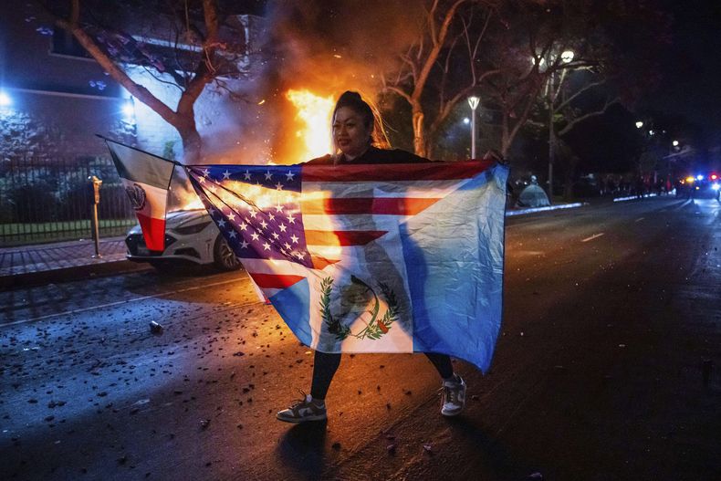 Una persona con una combinación de banderas camina frente a un auto incendiado durante las protestas por las redadas del gobierno de Trump en Los Ángeles, el lunes 9 de junio de 2025. (AP Foto/Ethan Swope)