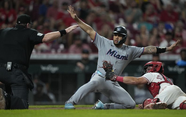 El receptor Tyler Stephenson, derecha, de los Rojos de Cincinnati, observa al umpire Chris Conroy, izquierda, mientras Agustín Ramírez, centro, de los Marlins de Miami llega a salvo al plato con un sencillo de Liam Hicks durante la quinta entrada del juego de béisbol de Grandes Ligas el lunes 7 de julio de 2025, en Cincinnati. (AP Foto/Carolyn Kaster)