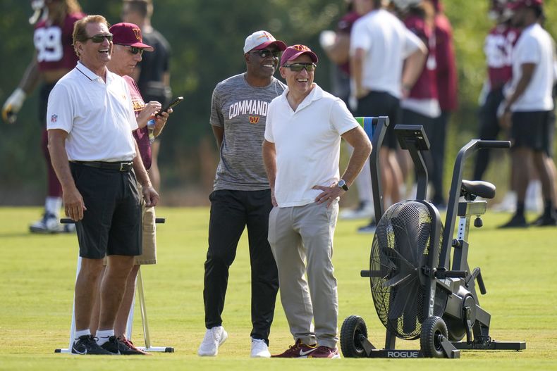Josh Harris, derecha, nuevo dueño de los Commanders de Washington, sonríe con el legendario Joe Theismann, izquierda, Mitchell Rales, y otros, durante el entrenamiento del equipo en las instalaciones, el miércoles 26 de julio de 2023, en Ashburn, Virginia. (AP Foto/Alex Brandon)