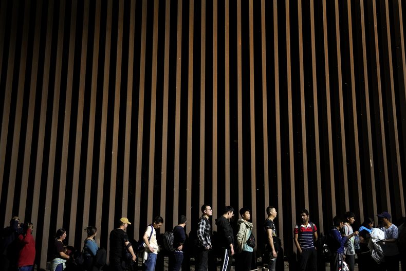 ARCHIVO - Personas formadas junto a un muro fronterizo mientras esperan solicitar asilo en Estados Unidos tras cruzar la frontera desde México, el martes 11 de julio de 2023, cerca de Yuma, Arizona. (AP Foto/Gregory Bull, Archivo)