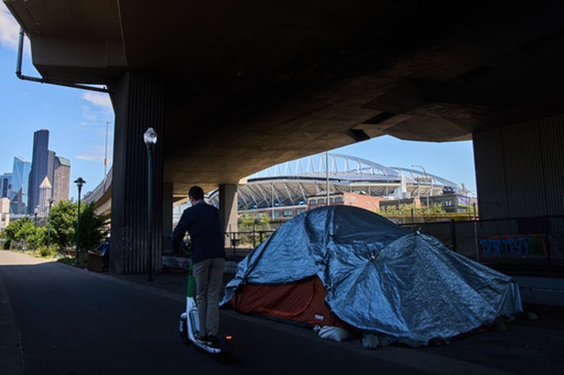 Un campamento de personas sin hogar debajo de un puente mientras a lo lejos se observa el Seattle Stadium y una persona cruza en un scooter antes del inicio de la Copa Mundial el miércoles 27 de mayo del 2026 en Seattle. (AP Foto/Lindsey Wasson)